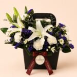 White lily and blue flower arrangement in a black handbag box